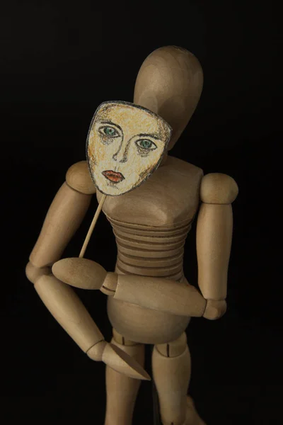 Dřevěná panenka na panty drží masku v rukou a pokrývá její tvář na černém pozadí — Stock fotografie