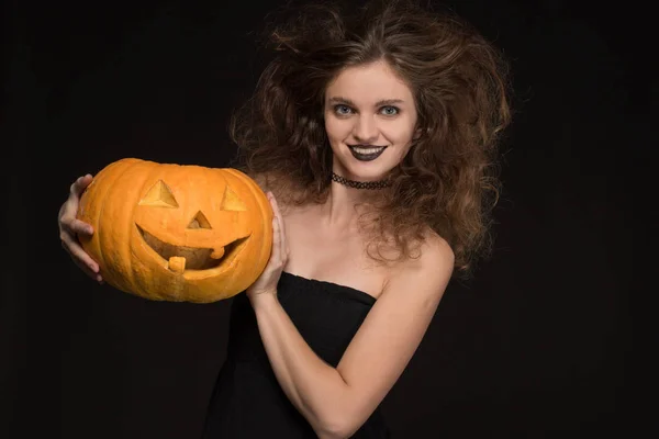 Mooi meisje met een make-up als een heks glimlacht en houdt een oranje pompoen op een zwarte achtergrond voor de vakantie van Halloween — Stockfoto
