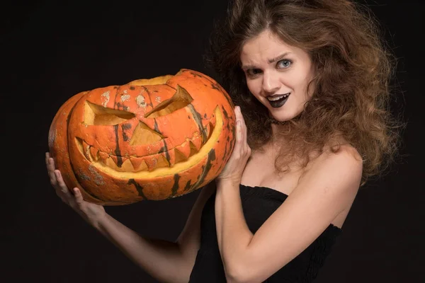 Mooi meisje met een make-up als een heks glimlacht en houdt een oranje pompoen op een zwarte achtergrond voor de vakantie van Halloween — Stockfoto
