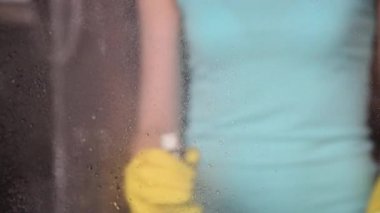 Genç bir ince kadın sarı eldiven su ve paçavra ve sünger ile bir sprey kullanarak bir evde pencereler yıkar