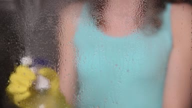 Genç bir ince kadın sarı eldiven su ve paçavra ve sünger ile bir sprey kullanarak bir evde pencereler yıkar