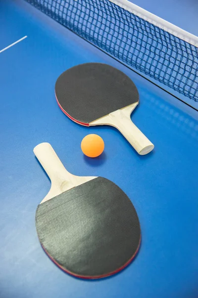 Orange ball for table tennis and two wooden rackets of red and black color on a blue table with a grid