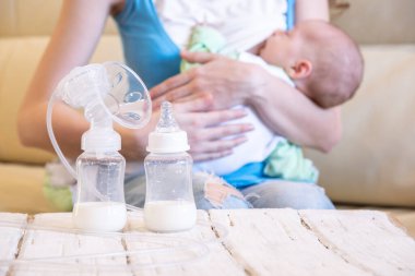 Genç bir anne bebeğini emziriyor ve elektrikli bir göğüs pompası önünde tahta bir masanın üzerinde duruyor.