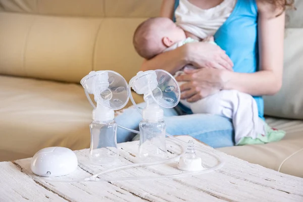 Genç bir anne bebeğini emziriyor ve elektrikli bir göğüs pompası önünde tahta bir masanın üzerinde duruyor.