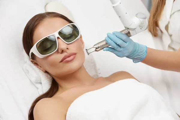 stock image beautician wearing gloves and holding laser near beautiful woman lying with protection glasses in beauty salon, woman receiving facial treatment in spa