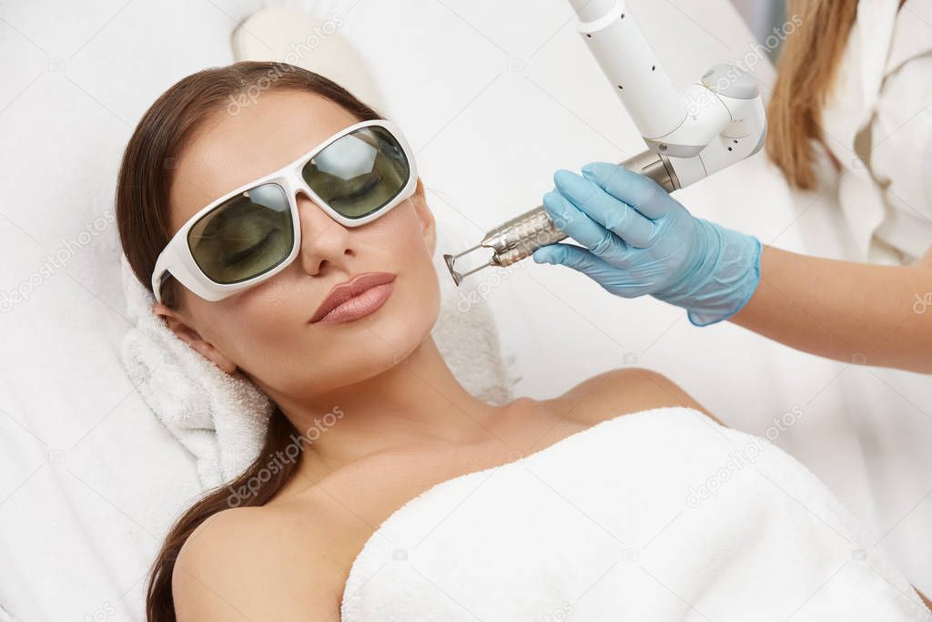 beautician wearing gloves and holding laser near beautiful woman lying with protection glasses in beauty salon, woman receiving facial treatment in spa