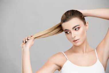 Güzel bir kadın sarı saçlarını yana doğru çekiyor ve gri renkte izole bir şekilde ona bakıyor.