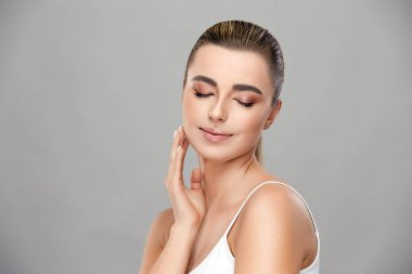 Gözleri kapalı gülümseyen ve gri arka planda yanağına dokunan açık makyajlı güzel bir kız.