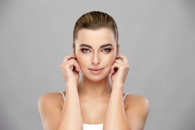 Güzel bayan kulaklarına dokunuyor ve gri arka planda kameraya bakıyor.