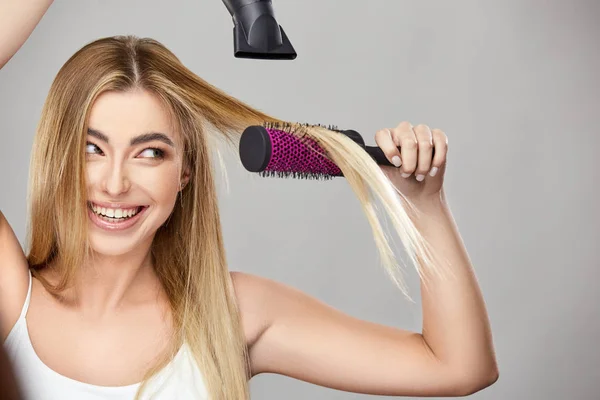 Pretty blond uses hair dryer for her healthy long hair and smiling — Stock Photo, Image