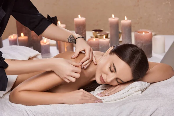 professional masseur doing shoulder massage for female customer in beauty salon with candles on background, young attractive girl in spa relaxing and having massage 