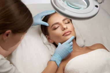 Güzellik kliniğinde, ışığın altında uzanmış güzel gözleri olan güzel bir kız ve güzellik uzmanı yüzünü eldivenlerle kontrol ediyor, güzel bir kadın güzellik salonunda cilt bakımı yaptırıyor.