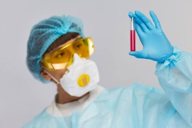 Bilim adamının kolunda kırmızı sıvı bulunan mavi eldiven ve koruyucu kostüm giymiş test tüpünün yakın çekimi.