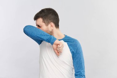 Mavi ve beyaz gömlekli sakallı adam, diğerlerini koronavirüsten korumak için dirseğine hapşırıyor.