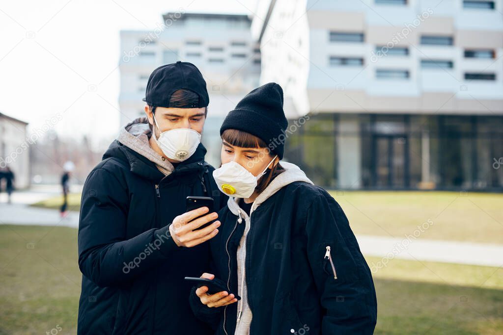 pretty couple in black clothes standing on the street with modern architecture in respiratory mask and showing news on phone, stylish man showing telepnone new to girl outdoor in masks