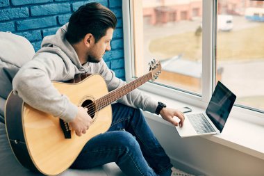 Gitarlı bir adam pencerede dizüstü bilgisayar kullanıyor modern dairede müzik aleti olan neşeli bir adam.