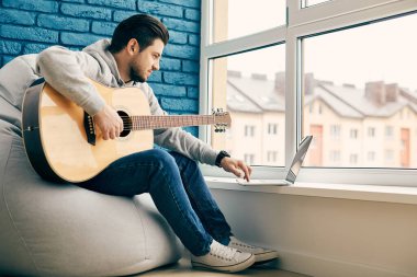 Yeni gitar öğrenen yakışıklı bir adam. Modern odada gitar çalıyor. Gri bomba koltuğunda oturan enstrümanı kullanıyor.