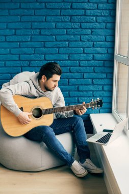 Gitarlı romantik bir adam evde laptopuyla tuğla duvarda pratik yapıyor. Dikey çekimde erkek, müzik aletini asgari seviyede tutuyor.