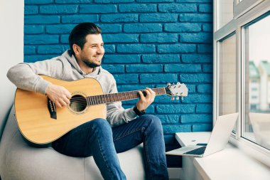 Mutlu bir adam evde dizüstü bilgisayarla gitar çalıyor. Evde müzik aleti olan yakışıklı bir erkek.