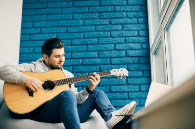 Gitar tutan erkeksi adam modern odada oturuyor mavi tuğlalardan ve pencerenin pervazında bacakları var.