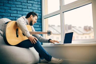 Neşeli bir adam bacaklarında gitarla ona gülümseyen bir dizüstü bilgisayar kullanıyor, balkonda enstrümanlı bir gitarist oturuyor ve bilgisayara gülüyor.