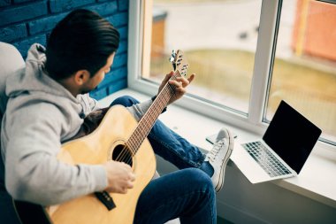 Pencere pervazında dizüstü bilgisayarla gitar çalan müzisyen ve mavi duvar
