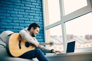 Modern dairede oturan ve dizüstü bilgisayar kullanan mutlu bir gitarist.