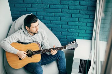 Modern odada gündelik giyinen gitarist mavi tuğlayla pencere pervazında pratik yapıyor, boşluğu kopyalıyor.