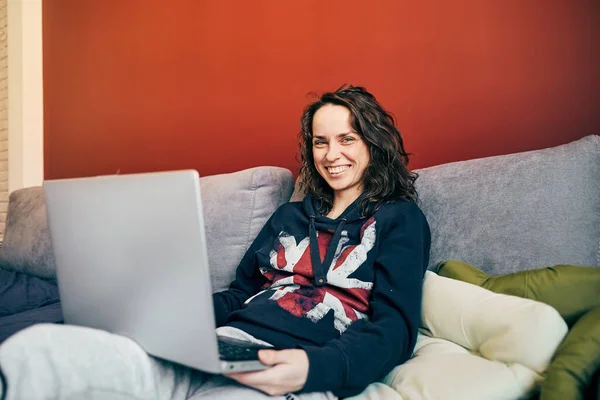 handsome woman sitting on couch with laptop and smiling to camera, lovely girl in british flag sweater working remotely