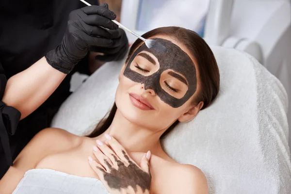 close-up of woman face with carbon mask and cosmetologist arm applying it, lovely girl receiving facial treatment by beautician