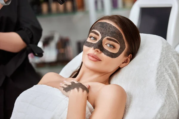 pretty woman with carbon mask on face lying in spa and looking to camera, beauty procedures and facial treatment