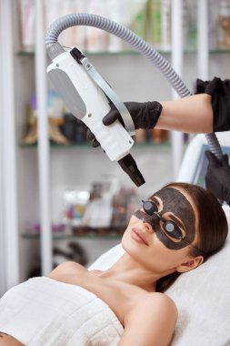 Yüzünde karbon maskesi olan güzel bir kız kaplıca salonunda kozmetik uzmanı tarafından siyah gözlüklerle lazer prosedürleri görüyor.