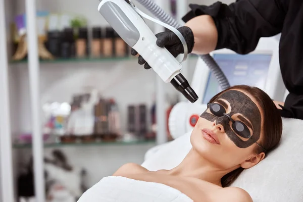 woman in spa with black carbon mask and cosmetologist with laser near face, copy space, lovely girl having rejuvenation by beautician