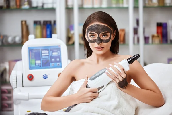 sexy girl with mask on face holding beauty aparat and looking to camera, beauty machine in arms of strong woman at spa salon
