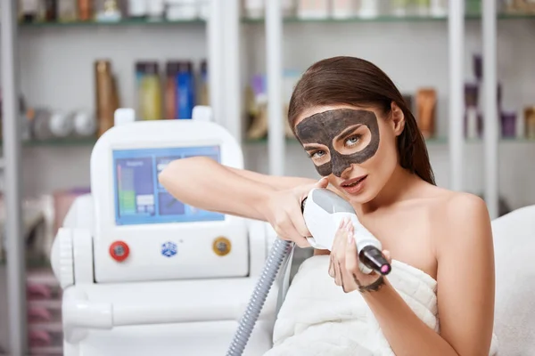 gorgeous woman with black carbon mask on face pointing laser gun to the camera at spa clinic, sexy chic shooting with beauty machine at beauty salon