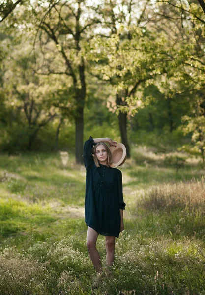 Ein hübsches Mädchen in einem schwarzen Kleid hält einen Strohhut — Stockfoto