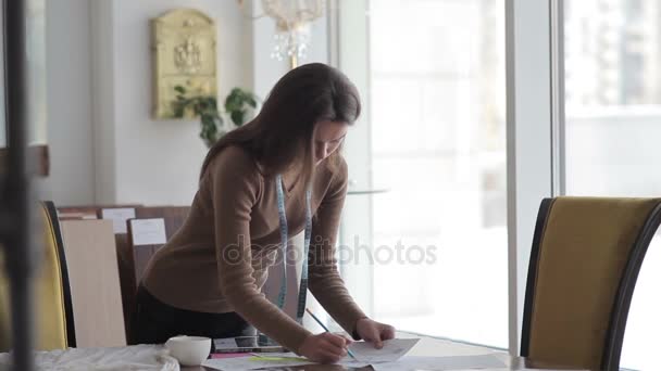 テーブルの近くに立って計算を行う若い女性 紙書類の山とテーブルの近く立って図面を作る労働者 首と鉛筆を手に巻尺で立っている女性 — ストック動画