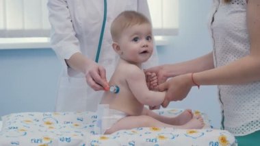 Yakın çekim. Küçük çocuk bebek bezi üzerinde oturur. Anne oğlu tarafından ellerini tutar. Doktor stetoskop ile çocuk gövdesini dinle. 