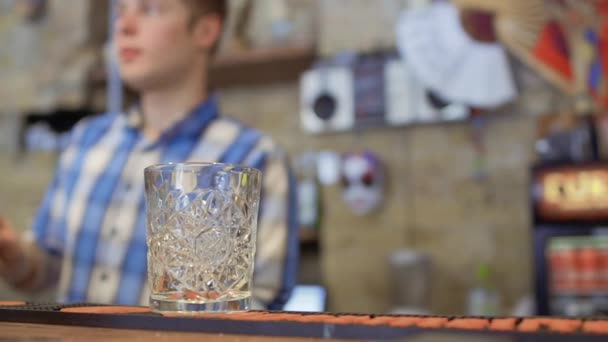Пустой стакан на барной стойке, бармен готовит коктейль на размытом фоне — стоковое видео