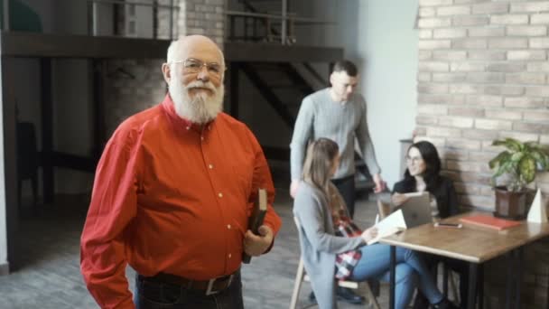 Reifer Mann mit Buch blickt auf junges Business-Team vor verschwommenem Hintergrund — Stockvideo