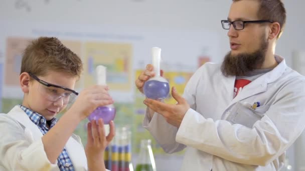 Учёный и его ученик держат в руках колбы с парящими красочными жидкостями — стоковое видео