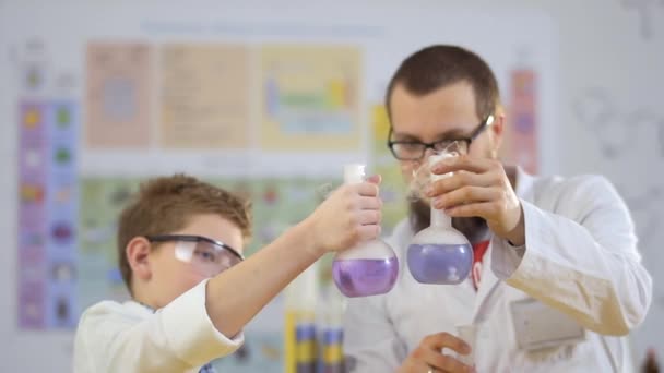 Laboratorium assistent met leerling kijkt stomen kleurrijke vloeistoffen in kolven — Stockvideo