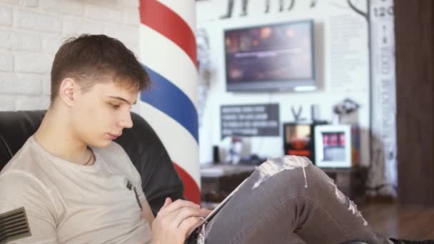 Jonge kerel maakt gebruik van de Tablet PC en wacht op zijn beurt in de barbershop — Stockvideo