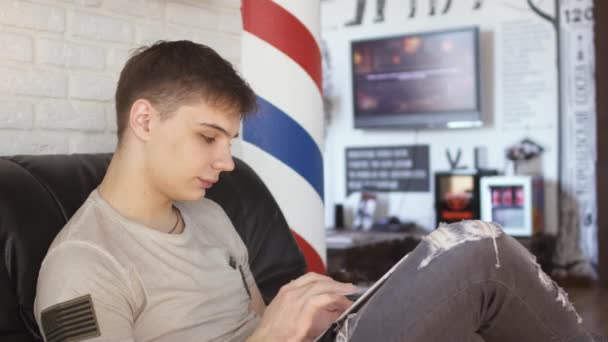 Knappe jongen maakt gebruik van de Tablet PC en wacht op zijn beurt in de barbershop — Stockvideo