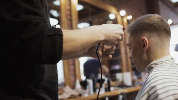 Peluquero corta el cabello con maquinilla de afeitar eléctrica a cliente joven — Vídeo de stock