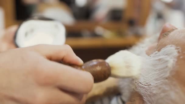 Stylist applies shaving cream on a gray beard of senior man — Stock Video