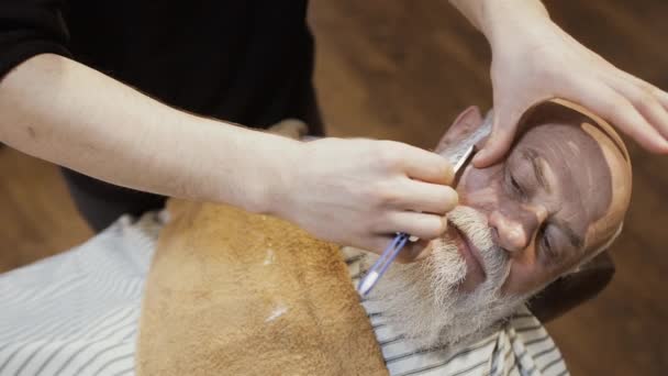 Barber shaves beard of senior man with blade in barbershop — Stock Video