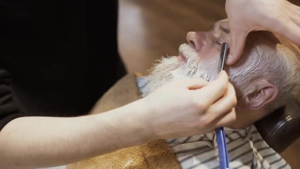 Friseur rasiert Gesicht eines älteren Mannes mit Klinge und Sahne — Stockvideo