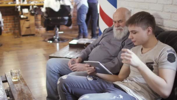 Молодой парень и зрелый мужчина используют цифровые планшеты и ждут своей очереди в парикмахерской — стоковое видео