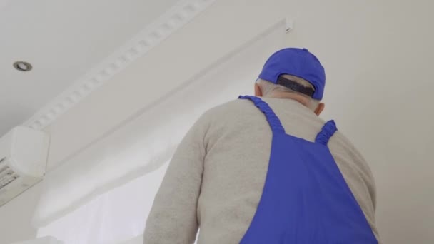 Äldre mannen i blå overaller reparera fatstening av rulljalusier — Stockvideo